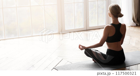 Sporty girl meditating in yoga studio, sitting in lotus pose, empty space 117500321