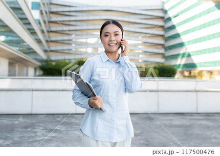 A young Asian businesswoman stands confidently outdoors, engaged in a phone conversation. She holds a clipboard in one hand and wears a light blue shirt. A young Asian businesswoman stands confidently outdoors, engaged in a phone conversation. She holds a clipboard in one hand and wears a light blue shirt. 117500476