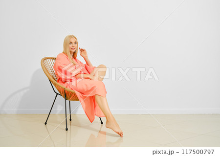 Cute woman in coral maxi dress sits in wicker chair in bright room 117500797