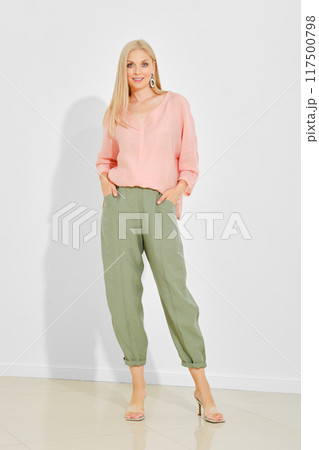 Stylish woman with smooth blonde hair wearing a pink blouse and green trousers 117500798