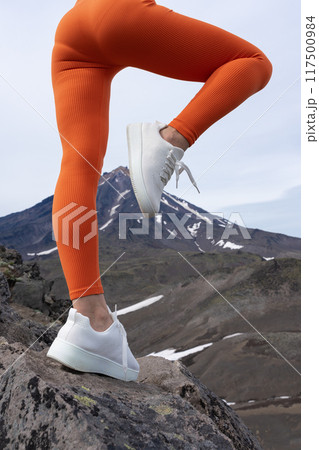 Close-up of fit woman's legs in orange workout leggings and sneakers doing outdoor fitness. Unrecognizable athlete practicing fitness in mountains and volcano. Sportswoman standing on one leg 117500984