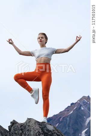 Flexible female yoga instructor balancing her body on one leg while doing yoga, standing on top of rocky mountain with volcano in background. Slim woman wears orange yoga leggings and sneakers Flexible female yoga instructor balancing her body on one leg while doing yoga, standing on top of rocky mountain with volcano in background. Slim woman wears orange yoga leggings and sneakers 117501015