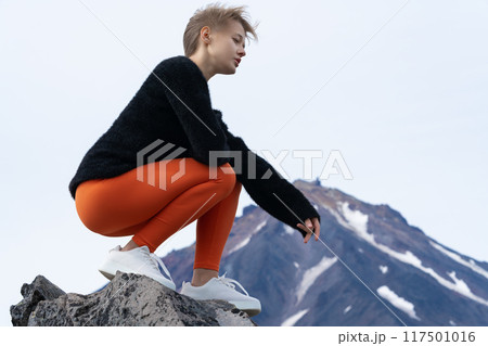 Woman tourist sits squatting on rocky outcrop in mountains during summer travel through mountainous area with volcano cone in background. Lonely woman enjoying moment of tranquility, looking at view 117501016