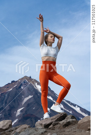 Happy and serene female athlete stands in mountains against backdrop of volcano cone. Beautiful slender blonde yoga instructor with short hair is dressed in t-shirt, orange yoga pants and trainers Happy and serene female athlete stands in mountains against backdrop of volcano cone. Beautiful slender blonde yoga instructor with short hair is dressed in t-shirt, orange yoga pants and trainers 117501025