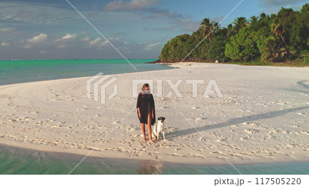 Young woman is enjoying a relaxing walk with her dog on a pristine white sand beach, with lush tropical vegetation and turquoise waters in the background 117505220