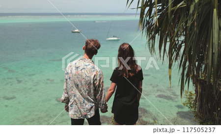 Young couple holding hands, admiring the ocean from a tropical cliff, with turquoise waters blending into the sky above Young couple holding hands, admiring the ocean from a tropical cliff, with turquoise waters blending into the sky above 117505237