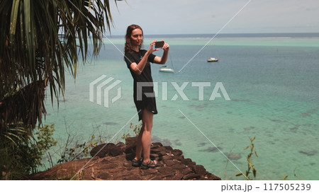 A woman stands on the shore, holding a camera up to take a picture of a boat gliding across the water. 117505239