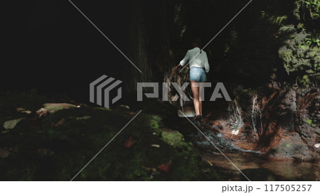 Female tourist is carefully walking on rocks in a cave, behind a small waterfall, discovering the beauty of tropical nature Female tourist is carefully walking on rocks in a cave, behind a small waterfall, discovering the beauty of tropical nature 117505257