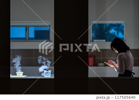 キッチンで夕食を作りながらスマホを操作する若い女性のイメージ素材 117505640