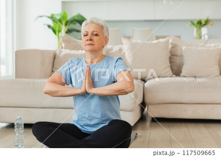 Yoga mindfulness meditation. Senior adult mature woman practicing yoga at home. Mid age old lady sitting in lotus pose on yoga mat meditating relaxing. Older middle aged woman doing breathing practice Yoga mindfulness meditation. Senior adult mature woman practicing yoga at home. Mid age old lady sitting in lotus pose on yoga mat meditating relaxing. Older middle aged woman doing breathing practice 117509665