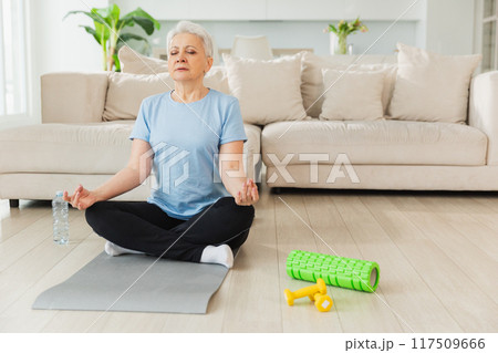 Yoga mindfulness meditation. Senior adult mature woman practicing yoga at home. Mid age old lady sitting in lotus pose on yoga mat meditating relaxing. Older middle aged woman doing breathing practice Yoga mindfulness meditation. Senior adult mature woman practicing yoga at home. Mid age old lady sitting in lotus pose on yoga mat meditating relaxing. Older middle aged woman doing breathing practice 117509666