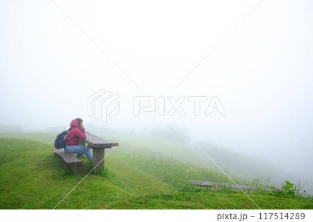 Asian young woman in red raincoat and bag sitting on wooden bench in green grass field and foggy with mist on mountain in rainy season. Travel in Asian Asian young woman in red raincoat and bag sitting on wooden bench in green grass field and foggy with mist on mountain in rainy season. Travel in Asian 117514289