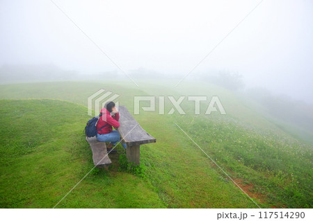 Asian young woman in red raincoat and bag sitting on wooden bench in green grass field and foggy with mist on mountain in rainy season. Travel in Asian 117514290