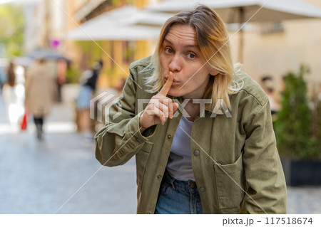 Young woman makes silence hush gesture sign asking do not tells gossip secret on urban city street Young woman makes silence hush gesture sign asking do not tells gossip secret on urban city street 117518674
