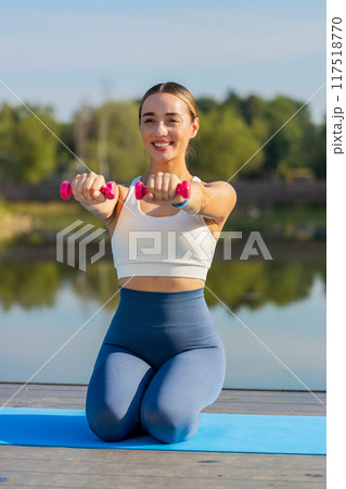 Sportive young woman exercising with dumbbells sitting on yoga mat near pond in park on sunny day 117518770