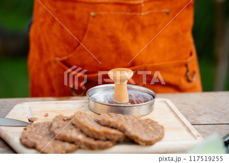Man in apron preparing meat for hamburgers, standing outdoors. Using burger patty press to shape groud meat. Man in apron preparing meat for hamburgers, standing outdoors. Using burger patty press to shape groud meat. 117519255