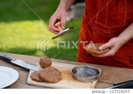 Man in apron preparing meat for hamburgers, standing outdoors. Using burger patty press to shape groud meat. 117519257