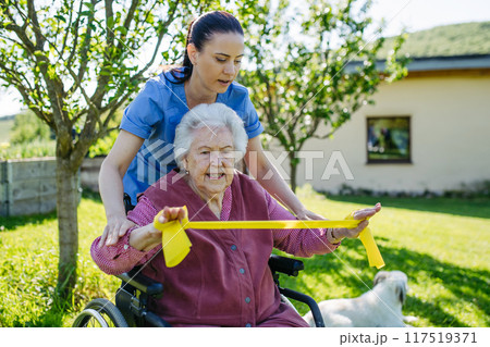 Female caregiver doing motorized exercises with senior woman in wheelchair. Nurse and elderly woman working out with resistance band. 117519371