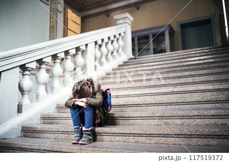 Portrait of bullied girl is crying on the steps in front of the school, waiting for her parents. Student girl with bag after day in elementary school. 117519372