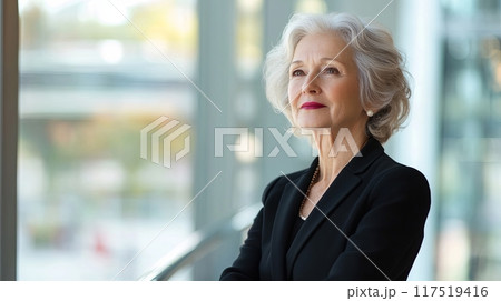 confident older woman dressed in professional attire, standing in a modern office space with large windows 117519416