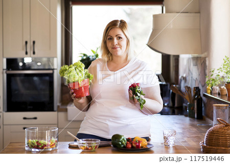Overweight woman in the kitchen, making homemade lunch. Choosing healthy food, making and eating vegetable salad. 117519446