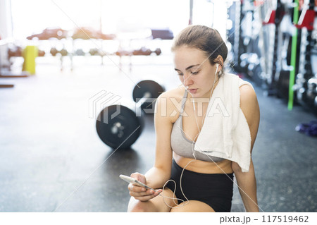Young fitness girl checking her performance on smatphone after hard workout in gym. Young fitness girl checking her performance on smatphone after hard workout in gym. 117519462