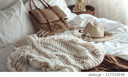 A soft, white knitted sweater and a stylish woven bag with leather straps rest on a neatly made bed 117519546