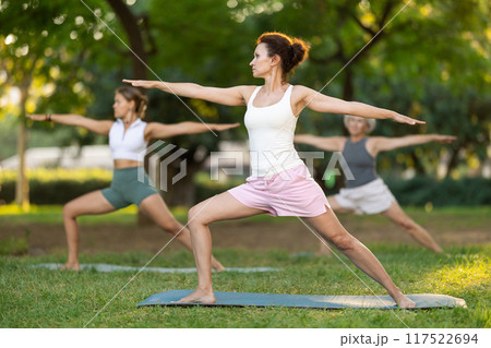 Happy woman standing yoga pose and making full body stretching workout with group people at park outdoor 117522694