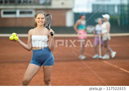 Cheerful girl with racket and tennis balls standing on outdoor court Cheerful girl with racket and tennis balls standing on outdoor court 117522815