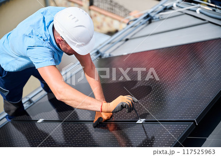 Worker building photovoltaic solar panel system on rooftop of house. Man engineer in helmets and gloves installing solar module with help of hex key outdoors. Renewable energy generation concept. Worker building photovoltaic solar panel system on rooftop of house. Man engineer in helmets and gloves installing solar module with help of hex key outdoors. Renewable energy generation concept. 117525963