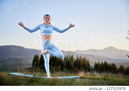 Woman practicing yoga and meditating outdoors in the mountains in a serene, natural setting. Female performing yoga pose on blue mat, with backdrop of beautiful mountain landscape at sunrise or sunset Woman practicing yoga and meditating outdoors in the mountains in a serene, natural setting. Female performing yoga pose on blue mat, with backdrop of beautiful mountain landscape at sunrise or sunset 117525976