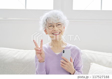 スマートフォンを持ってピースサインするシニア女性　リビングルーム 117527188