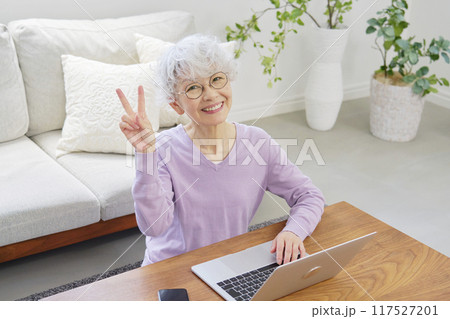 ノートパソコンの前でピースサインするシニア女性　リビングルーム 117527201