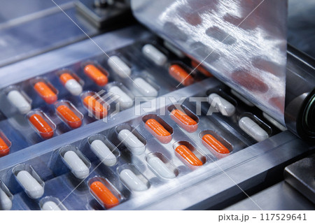 automated medicine packaging machine for orange and white capsules. Pharmaceutical production line with blister packs. Manufacturing and healthcare concept. Close-up shot. 117529641