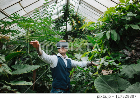 Handsome businessman standing in botanical garden, in the middle of lush green foliage, using VR headset. Handsome businessman standing in botanical garden, in the middle of lush green foliage, using VR headset. 117529887