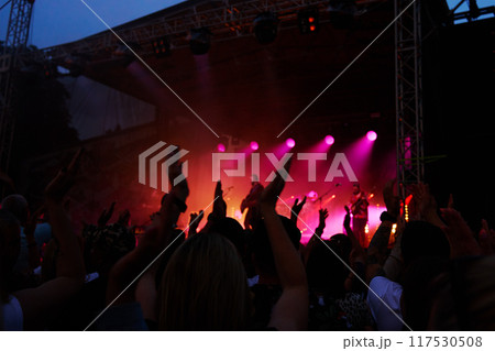 Silhouettes of crowd at concert in front of stage with bright spotlights. People enjoy live music at the band's performance. Concert hall with musicians on stage and fans during music festival Silhouettes of crowd at concert in front of stage with bright spotlights. People enjoy live music at the band's performance. Concert hall with musicians on stage and fans during music festival 117530508