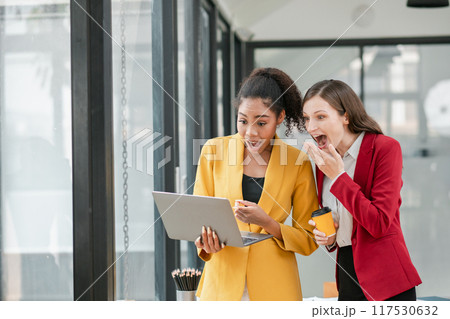 Two professional women in business attire express excitement while looking at a laptop in a contemporary office setting. 117530632