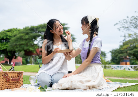 Two cheerful and pretty Asian women are enjoying picnic together in a green park on a bright day. 117531149