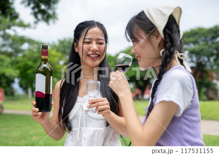 Two happy Asian women are having a picnic together in a green park, drinking red wine. 117531155