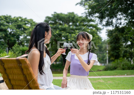 Two happy, charming young Asian women are enjoying red wine together during a picnic in a green park Two happy, charming young Asian women are enjoying red wine together during a picnic in a green park 117531160