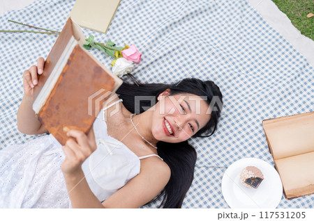 A top view of a positive, lovely Asian woman reading a book and lying on a picnic blanket. 117531205