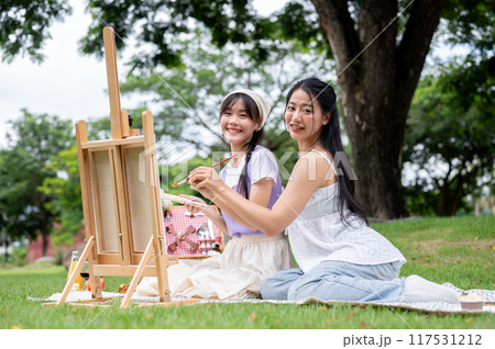Two pretty Asian women are sitting together on a picnic blanket with a canvas easel in a park. Two pretty Asian women are sitting together on a picnic blanket with a canvas easel in a park. 117531212