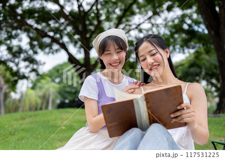 Two beautiful and positive Asian friends are reading a book together while picnicking in a park. Two beautiful and positive Asian friends are reading a book together while picnicking in a park. 117531225