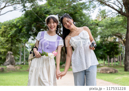 Two lovely Asian women are holding hands, strolling through a park, and smiling at the camera. Two lovely Asian women are holding hands, strolling through a park, and smiling at the camera. 117531228