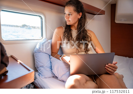 Young woman freelancer using laptop and looking out the window in the cabin of a yacht during sailing in sea. Yacht life concept Young woman freelancer using laptop and looking out the window in the cabin of a yacht during sailing in sea. Yacht life concept 117532011