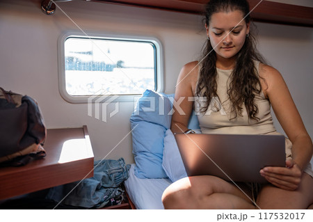 Young woman freelancer using laptop near window in the cabin of a yacht during sailing in sea. Yacht life concept 117532017