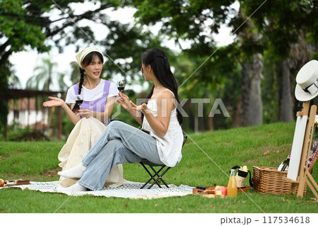 Young female friends drinking wine and enjoys a picnic on a blanket in a park Young female friends drinking wine and enjoys a picnic on a blanket in a park 117534618