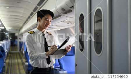 Smiling asian pilot standing inside an airplane, holding clipboard and reviewing or completing some paperwork Smiling asian pilot standing inside an airplane, holding clipboard and reviewing or completing some paperwork 117534660