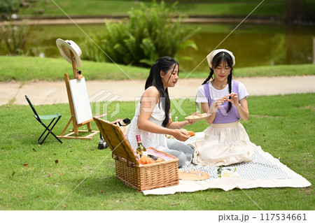 Relaxed young women friends sitting on a blanket in a park, eating fruit and drinking wine Relaxed young women friends sitting on a blanket in a park, eating fruit and drinking wine 117534661