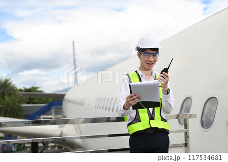 Young asian man in a yellow vest with walkie talkie standing on a metal railing next to an airplane 117534681